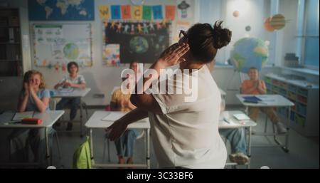 Fröhliche, abwechslungsreiche Kinder sitzen an Schreibtischen, klatschen die Hände, interagieren mit dem Lehrer während der dynamischen Gamified-Aktivität. Lehrerin, die Grundschüler interaktiven Unterricht gibt. Stockfoto