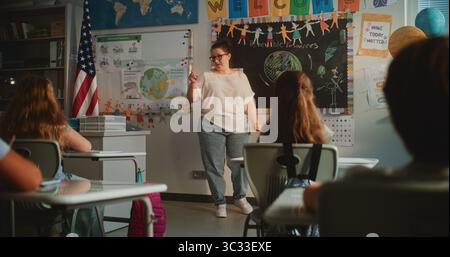 Lehrerin, die Vorlesungen über erneuerbare Energien für Grundschüler durchführt. Intelligente, vielfältige Kinder sitzen an Schreibtischen im modernen, farbenfrohen Klassenzimmer, studieren Ökologie, hören dem Lehrer zu. Stockfoto