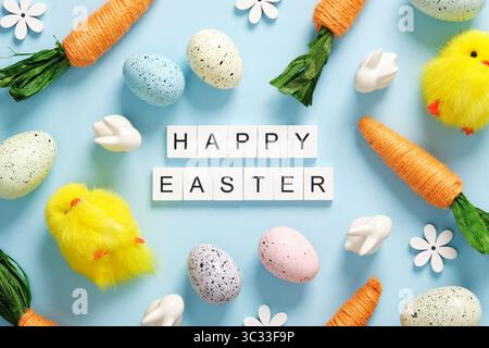 Text Happy Easter aus Holzblöcken mit farbigen Eiern, Hasen, Karotten und Hühnern auf blauem Hintergrund Stockfoto