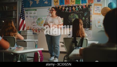 Lehrerin, die Vorlesungen über erneuerbare Energien für Grundschüler durchführt. Intelligente, vielfältige Kinder sitzen an Schreibtischen im modernen, farbenfrohen Klassenzimmer, studieren Ökologie, hören dem Lehrer zu. Stockfoto