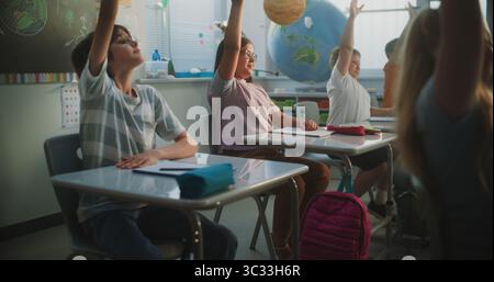 Grundschüler sitzen an den Schreibtischen, hören den Vorträgen des Lehrers zu und heben die Hände, um die richtige Antwort zu geben. Team von Smart Diversity Kids studiert Wissenschaft, Geographie im modernen Klassenzimmer. Stockfoto