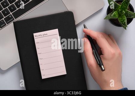 Die Hand des Mannes hält einen Stift. Daneben ist ein leerer Wochenplan, ein Notizbuch, eine Pflanze. Stockfoto