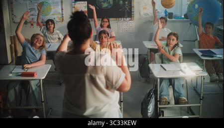Rückansicht der Lehrerin, die Grundschüler unterrichtet, Fragen stellt. Ein Team von Smart Diversity Kids sitzt an Schreibtischen, hebt die Hände, um Antworten zu geben, und studiert im modernen Klassenzimmer Stockfoto