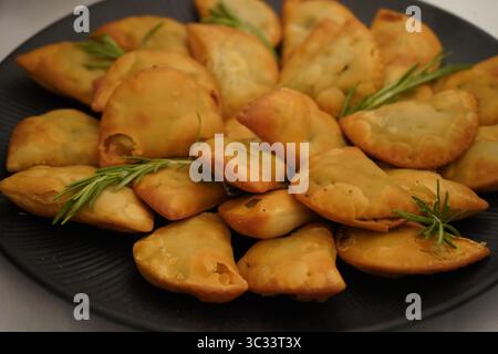 Goldbraune, halbmondförmige Grüns- und Kräuterkuchen sind auf einem dunklen Teller angeordnet. Garniert sind sie mit frischen Rosmarin-Zweigen Stockfoto