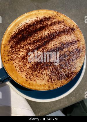 Blick von oben auf einen Cappuccino, der in einer Tasse mit dunklem Petrol auf einer hellen Untertasse serviert wird, die großzügig mit einem dunklen, körnigen Pulver bestreut ist. Stockfoto