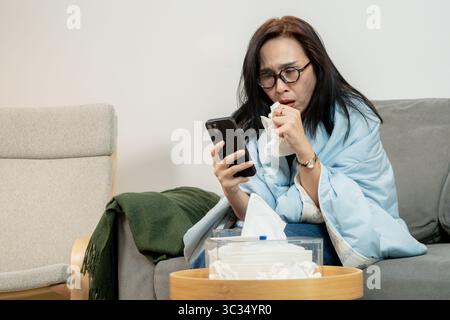 Eine junge Frau mit Schüttelfrost und Fieber sitzt in einer Decke auf der Couch zu Hause und nimmt Medikamente zur Linderung und Selbstpflege ein. Stockfoto