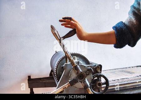 Handbetätigte manuelle Druckmaschine des Künstlers im traditionellen Druckstudio. Hand mit Flecken in der Druckertinte, die den Kurbelgriff dreht Stockfoto