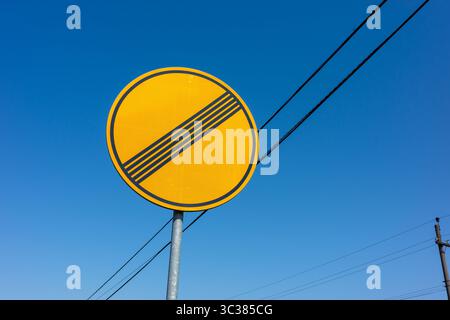 Ein Straßenschild, das das Ende einer Einschränkung anzeigt, wird vor einem klaren, hellblauen Himmel angebracht. Elektrische Drähte kreuzen die Szene subtil. Stockfoto