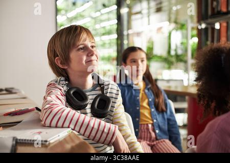 Kaukasischer Junge mit Kopfhörern am Schreibtisch, lächelnd und zur Seite schauend, multiethnische Gruppe von Kindern, die gemeinsam in modernen Klassenzimmern studieren, Notizbücher und Zubehör auf dem Tisch Stockfoto