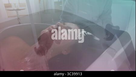 Liegende Frau spürt, wie Wasser aus dem Wasserhahn über die Stirn im Spa-Raum fließt, mit Badewanne und Chromhähnen Stockfoto
