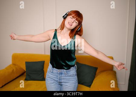 Frau mit großen Kopfhörern schaltet die Playlist ein hört Musik mit geschlossenen Augen, singt und tanzt im Wohnzimmer des Hauses. Fröhliches Mädchen genießt Musik, Entspannung, Ruhe zu Hause Stockfoto