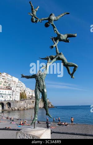 Amalfi, Italien-4. November 2024; Bronzeskulptur La Familia in Flight des Künstlers Andrea Roggi am Strand mit Sonnenbaden und weißen Häusern Stockfoto