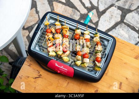 Eine Reihe hausgemachter Hühnchen- und Gemüsespieße, die auf einem elektrischen Moulinex-Grill grillen, auf einem Holztisch im Freien. Ein gesundes und farbenfrohes Di Stockfoto