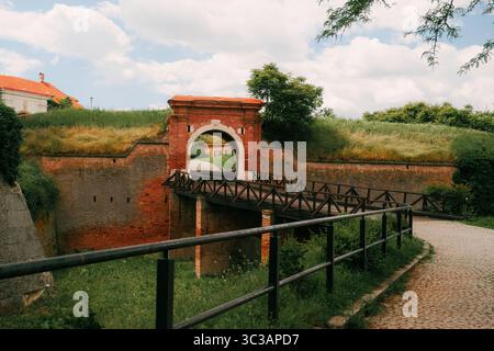 Historisches Tor aus rotem Backstein und hölzerne Brücke in der Festung Petrovaradin in Novi Sad, Serbien, umgeben von Grün und Kopfsteinpflasterweg Stockfoto