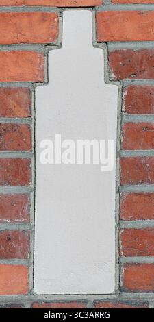 Nahaufnahme einer einzelnen vertikalen Putzeinlage, eingebettet in eine rote Ziegelwand. Die Form ähnelt einer stilisierten architektonischen Säulen- oder Flaschensilhouette. Stockfoto