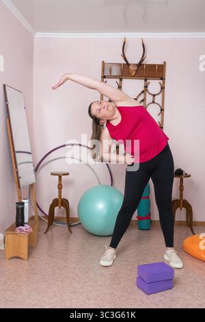 Frau mittleren Alters beugt sich mit erhobenem Arm während des Fitnesstrainings zu Hause. Rundumsicht einer 44-jährigen Kurvenfrau in Rot Stockfoto
