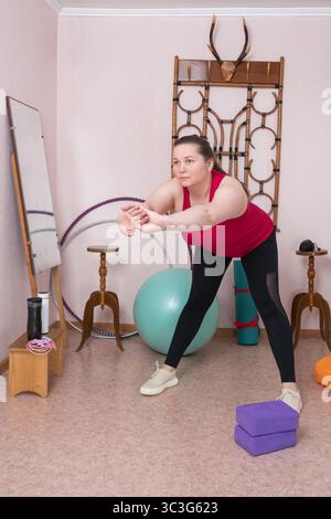 Frau mittleren Alters macht beim Fitnesstraining zu Hause Vorwärtsbeugung. Rundumsicht einer 44-jährigen Frau mit Sportbekleidung - rotes Tanktop, Stockfoto