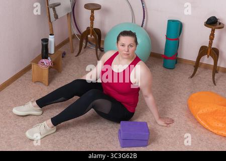 Erwachsene Fitness-Frau sitzt auf dem Boden und entspannt sich während des Pilates-Trainings zu Hause im Innenraum. Sportliche Frau ist Generation X im Sport gekleidet Stockfoto