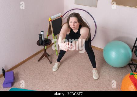 Die reife Frau führt während des häuslichen Fitness-Regimes eine Vorwärtsbeugung durch. Ganzkörperaufnahme einer erwachsenen Fitnessfrau kaukasischer Ethnizität in Bla Stockfoto