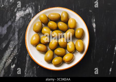 Grüne Oliven in einer Schüssel. Mediterraner Snack auf schwarzer Marmoroberfläche. Gesundes Vorspeisenkonzept. Bio-Obst-Nahaufnahme. Vegetarische Naturkost. Stockfoto
