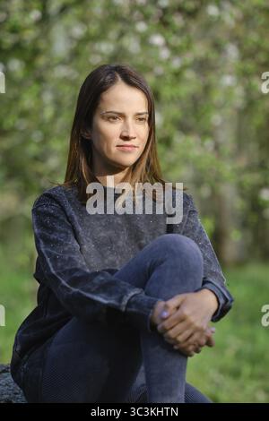 Eine Frau sitzt ruhig in einem Außenbereich, umgeben von üppigem Grün und sanftem Licht. Sie wirkt besinnlich, während sie im Kreuz auf einem Felsen sitzt Stockfoto