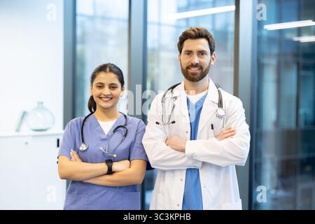 Zwei medizinische Fachkräfte, ein Arzt und eine Krankenschwester, stehen in einem modernen medizinischen Umfeld nebeneinander und lächelnd mit gekreuzten Armen. Stockfoto