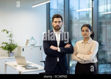 Ein professionelles Geschäftsteam, ein Mann und eine Frau, stehen selbstbewusst in einem Büro, mit überkreuzten Armen und Blick direkt in die Kamera. Stockfoto