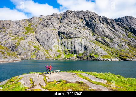 NUSFJORD VILLAGE, NORWEGEN - 19. Juni 2025: Zwei Wandererinnen mit Rucksack machen Fotos von Nusfjord Dorf und Fischerhafen auf Flakstadoya Island Stockfoto