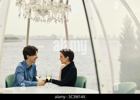 Kaukasischer junger Mann und kaukasische junge Frau sitzen an einem runden Tisch am Ufer, halten Hände und lächeln einander an, während sie Wein in elegantem Ambiente trinken Stockfoto