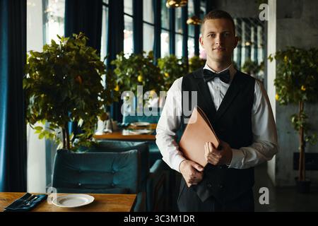 Porträt eines jungen Erwachsenen kaukasischen Mannes, der im Restaurant steht, Menüordner hält, mit formeller Weste und Fliege, der in die Kamera blickt, mit neutralem Ausdruck im Inneren Stockfoto