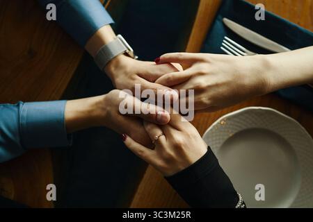 Kaukasischer Mann und kaukasische Frau, die Hände über einen Holztisch halten, beide mit langen Ärmeln, Frau mit Ring, Hände, die sanft an leeren Teller und Besteck klammern Stockfoto