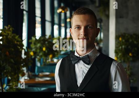 Porträt eines jungen Erwachsenen kaukasischen Mannes mit Fliege, der im Restaurant steht und die Kamera mit neutralem Ausdruck betrachtet, Inneneinrichtung mit verschwommenen Tischen und sichtbaren Pflanzen Stockfoto