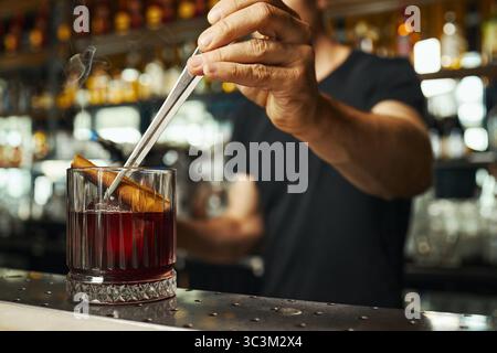 Kaukasischer junger Erwachsener, der Cocktail mit Zimtstab im Glas mit Metall-Barlöffel rührt, steht hinter der Bar im Restaurant mit Flaschenregalen im Hintergrund Stockfoto