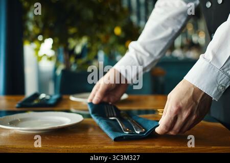 Kaukasischer junger Erwachsener, der Besteck und Serviette auf dem Restauranttisch arrangiert, Hände sichtbar, um Utensilien neben leeren Tellern zu platzieren und die Speisesaal für Gäste vorzubereiten Stockfoto