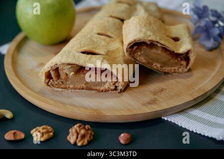 Köstlich gebackener Apfelstrudel auf einem Holzteller mit frischem grünem Apfel und verschiedenen Nüssen perfekt für ein leckeres Erlebnis Stockfoto