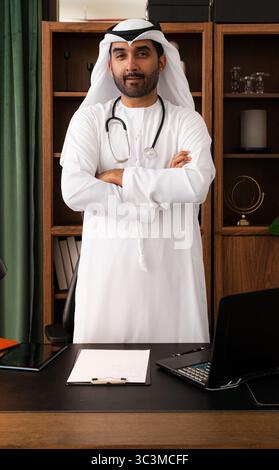 Nahöstlicher Arzt mit emirati-Kandura in der Praxis - zuversichtlicher nahöstlicher Arzt in traditioneller Kandura und Stethoskop. Exu Stockfoto