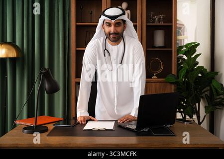Arzt aus dem Nahen Osten trägt emirat-Kandura im Büro - emirat-Arzt trägt traditionelle Kleidung und steht selbstbewusst an seinem Schreibtisch Stockfoto