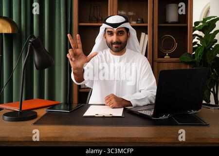 Geschäftsmann aus dem Mittleren Osten mit emirat Kandura im Büro - Geschäftsmann aus emirat in traditionellem Kandura, der an einem Schreibtisch sitzt und durch Gesten bewegt Stockfoto