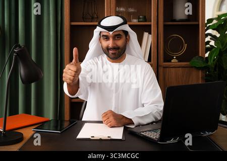 Geschäftsmann aus dem Nahen Osten mit emirat Kandura im Büro - erfolgreicher Unternehmer in traditionellem weißem Kandura am Schreibtisch mit l Stockfoto