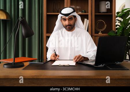 Geschäftsmann aus dem Mittleren Osten mit emirat Kandura, der im Büro arbeitet - Geschäftsmann aus emirat in traditionellem Kandura, der fleißig an seinem Schreibtisch arbeitet. Stockfoto