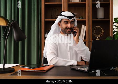 Geschäftsmann aus dem Nahen Osten trägt emirat Kandura, der im Büro arbeitet - Geschäftsmann aus emirat in traditionellem Kandura lächelt während er über das Telefon spricht Stockfoto