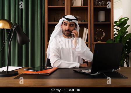 Geschäftsmann aus dem Mittleren Osten mit emirat Kandura, der im Büro arbeitet - Geschäftsmann aus emirat in traditionellem Kandura, der an einem modernen Schreibtisch sitzt. Stockfoto