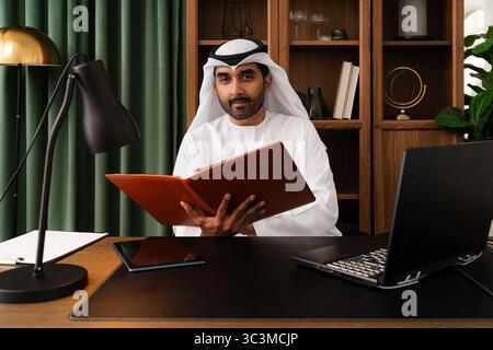 Geschäftsmann aus dem Mittleren Osten mit emirat Kandura, der im Büro arbeitet - Geschäftsmann aus emirat in traditionellem Kandura, der an einem modernen Schreibtisch sitzt. Stockfoto