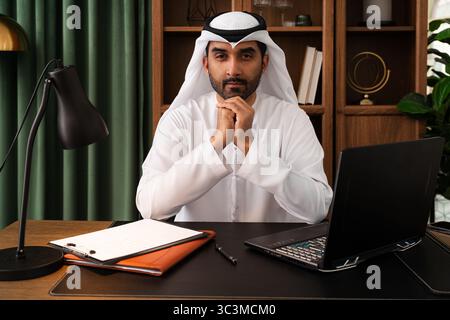 Geschäftsmann aus dem Mittleren Osten mit emirat Kandura, der im Büro arbeitet - zuversichtlicher emirat-Geschäftsmann in traditionellem Kandura, der in einem modernen von sitzt Stockfoto