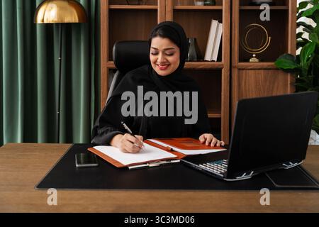 Ärztin oder Krankenschwester aus dem Mittleren Osten, die Abaya trägt und im Büro arbeitet - selbstbewusste Geschäftsfrau aus dem Mittleren Osten in Abaya und Hijab, die Notizen schreibt Stockfoto