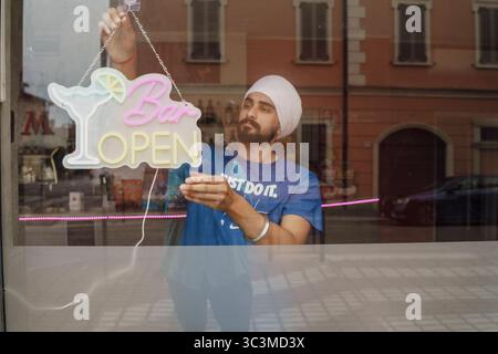 Cremona, Italien - 25. Juli 2025 ein Mann hängt ein offenes Schild im Fenster der Bar und begrüßt Gäste, die Getränke und Unterhaltung genießen können Stockfoto