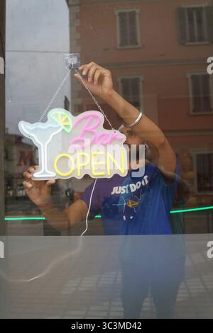 Cremona, Italien - 25. Juli 2025 ein Mann hängt ein offenes Neonschild im Fenster einer Bar mit einem Cocktailglas und einer Limettenscheibe Stockfoto