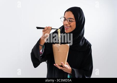 Die arabische Frau trägt traditionelle schwarze Abaya und Hijab und isst mit Essstäbchen eine köstliche Nudelmahlzeit, die Genuss und kulturelle Fusion ausdrückt Stockfoto