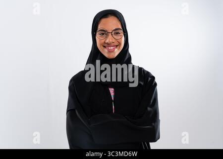 Studio-Porträt einer jungen, lächelnden arabischen Frau, die eine traditionelle schwarze Abaya und einen Hijab mit überkreuzten Armen trägt und muslimische Kultur und Mode repräsentiert Stockfoto