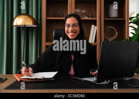 Geschäftsfrau aus dem Nahen Osten, die im Büro arbeitet - lächelnde Geschäftsfrau, die traditionelle Abaya trägt, arbeitet mit Laptop und nimmt Notiz Stockfoto